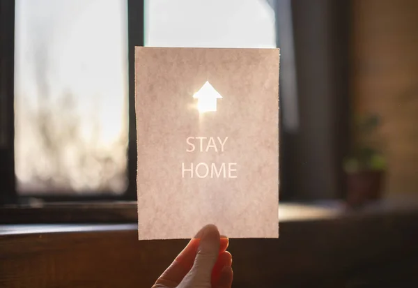 Hoja blanca de papel con un letrero de casa brillante y palabras Quédese en casa en una mano — Foto de Stock