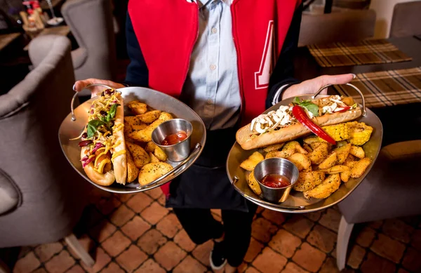 Číšník s dvěma talíři s velkým párkem a hranolkama. Restaurace. — Stock fotografie