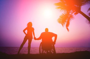 Disabled man in a wheelchair with his wife on the beach. Silhouettes at sunset