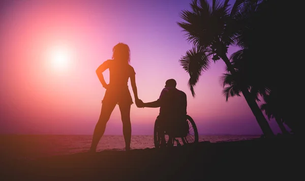 Disabled man in a wheelchair with his wife on the beach. Silhouettes at sunset — 스톡 사진