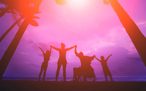 Hombre discapacitado en silla de ruedas con su familia en la playa. Siluetas al atardecer — Foto de Stock