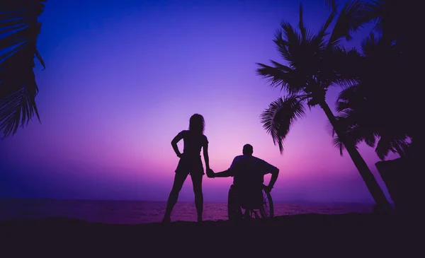 Disabled man in a wheelchair with his wife on the beach. Silhouettes at sunset — Stockfoto