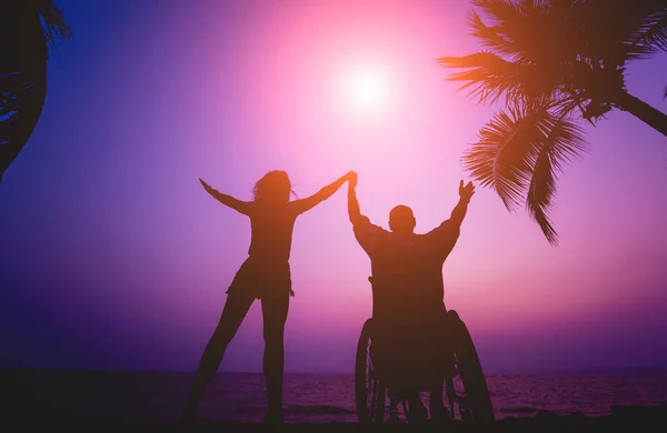 Hombre discapacitado en silla de ruedas con su esposa en la playa. Siluetas al atardecer — Foto de Stock