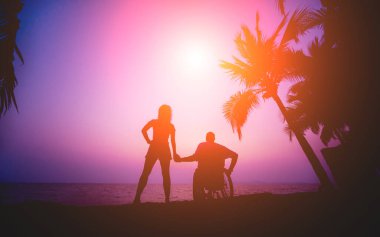 Disabled man in a wheelchair with his wife on the beach. Silhouettes at sunset