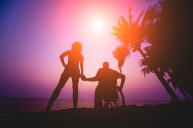 Disabled man in a wheelchair with his wife on the beach. Silhouettes at sunset