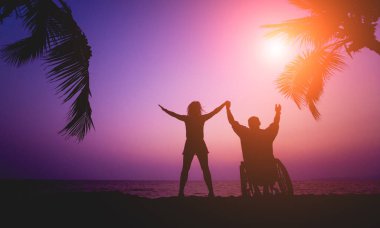 Disabled man in a wheelchair with his wife on the beach. Silhouettes at sunset