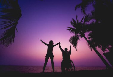 Disabled man in a wheelchair with his wife on the beach. Silhouettes at sunset