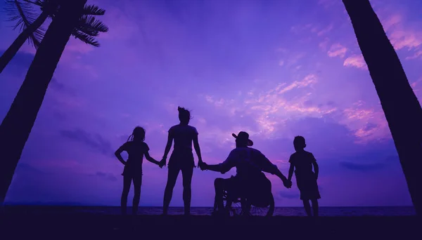 Hombre discapacitado en silla de ruedas con su familia en la playa. Siluetas al atardecer — Foto de Stock