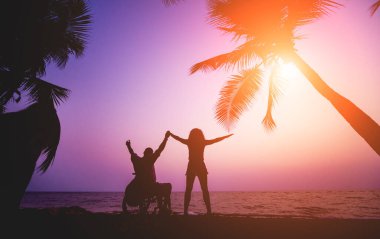Disabled man in a wheelchair with his wife on the beach. Silhouettes at sunset