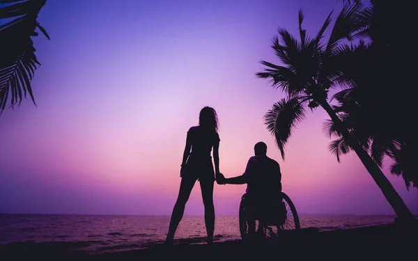 Инвалид в инвалидном кресле с женой на пляже. Силуэты на закате — стоковое фото