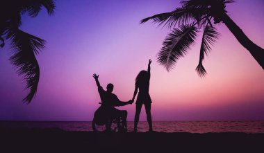 Disabled man in a wheelchair with his wife on the beach. Silhouettes at sunset