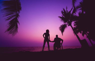 Disabled man in a wheelchair with his wife on the beach. Silhouettes at sunset