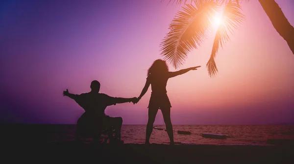 Disabled man in a wheelchair with his wife on the beach. Silhouettes at sunset — 스톡 사진