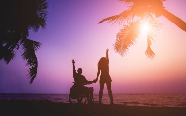 Disabled man in a wheelchair with his wife on the beach. Silhouettes at sunset