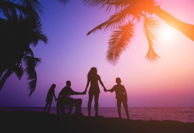 Disabled man in a wheelchair with his family on the beach. Silhouettes at sunset