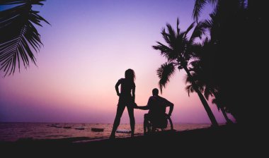 Disabled man in a wheelchair with his wife on the beach. Silhouettes at sunset