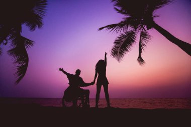 Disabled man in a wheelchair with his wife on the beach. Silhouettes at sunset