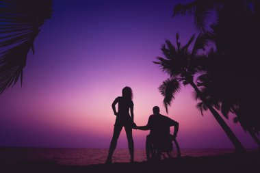 Disabled man in a wheelchair with his wife on the beach. Silhouettes at sunset