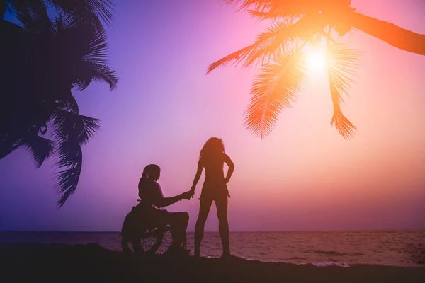 Инвалид в инвалидном кресле с женой на пляже. Силуэты на закате — стоковое фото