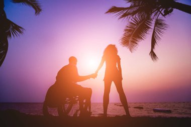 Disabled man in a wheelchair with his wife on the beach. Silhouettes at sunset