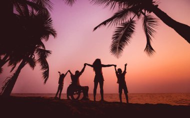 Disabled man in a wheelchair with his family on the beach. Silhouettes at sunset