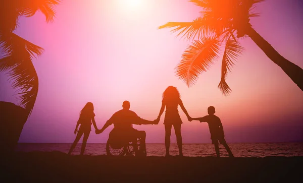 Um homem deficiente numa cadeira de rodas com a família na praia. Silhuetas ao pôr-do-sol — Fotografia de Stock