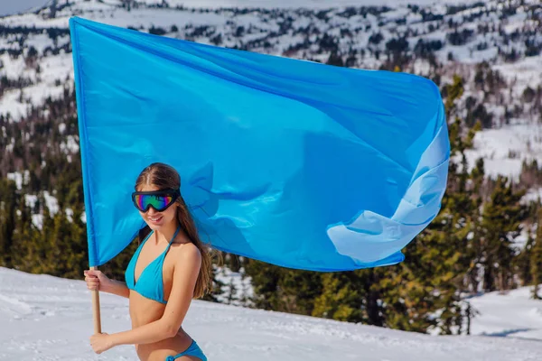 Hermosa mujer joven en traje de baño azul con gran bandera azul en la pendiente de invierno —  Fotos de Stock