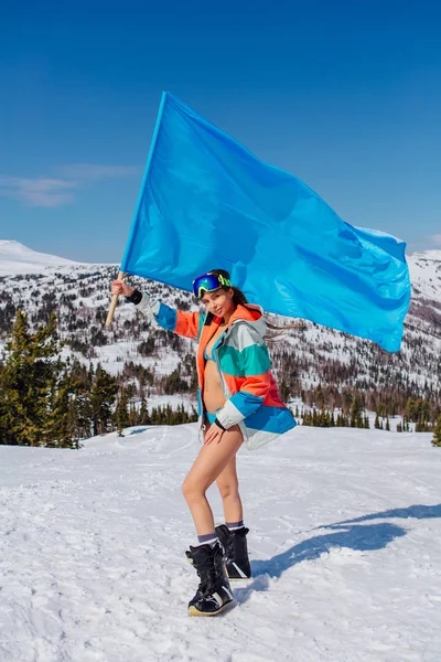 Vacker ung kvinna i blå baddräkt med stora blå flagg på sluttningen vinter — Stockfoto