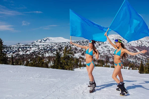 Två vackra unga kvinnor i blå baddräkter med stora blå flaggor på sluttningen vinter — Stockfoto