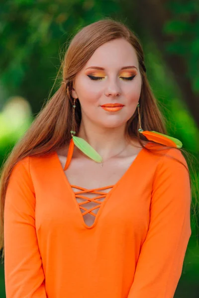 夏の公園で長い羽のイヤリングとオレンジのドレスで美しいロマンチックな赤い髪の少女 — ストック写真