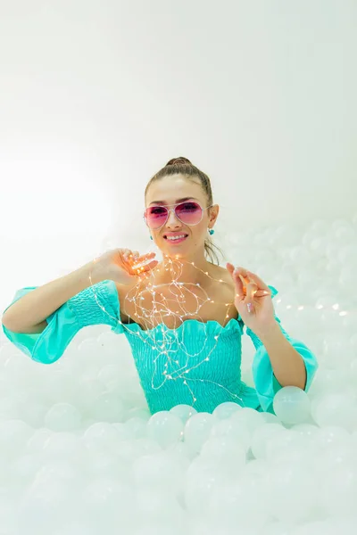 Feliz hermosa mujer yace rodeada de bolas de plástico blanco con guirnalda — Foto de Stock