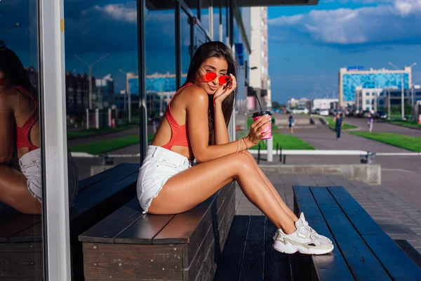 Elegante feliz jovem morena mulher vestindo branco shorts e tênis segurando rosa xícara de café para ir sentado ao lado de café . — Fotografia de Stock