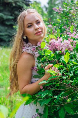 Leylak çalıyanında duran güzel beyaz elbise giyen büyüleyici sarışın bir kadın portresi.