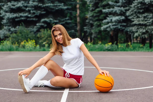 Gadis muda cantik berpakaian putih t-shirt, celana pendek dan sepatu olahraga, duduk di lapangan basket dengan bola . — Stok Foto