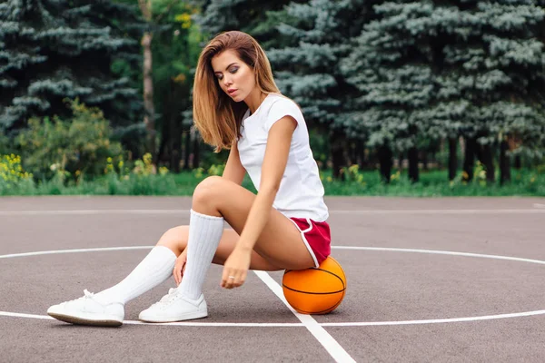 Gadis muda cantik berpakaian putih t-shirt, celana pendek dan sepatu olahraga, duduk di lapangan basket pada bola . — Stok Foto