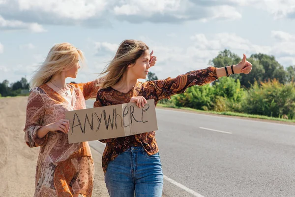 Two beautiful girls hitchhiking and vote with a sign ANYWHERE on road. Copy space. — Stock Photo, Image