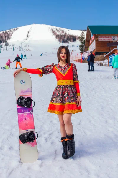 Sheregesh, Kemerovo region, Russia - April 12, 2019: Young happy pretty woman dressed in ethnic dress holding snowboard on snow slope. — Stock Photo, Image