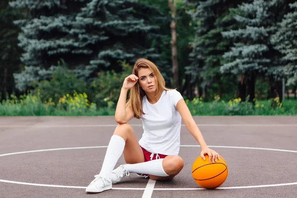 美丽的年轻女孩穿着白色T恤,短裤和运动鞋,坐在篮球场上与球. — 图库照片