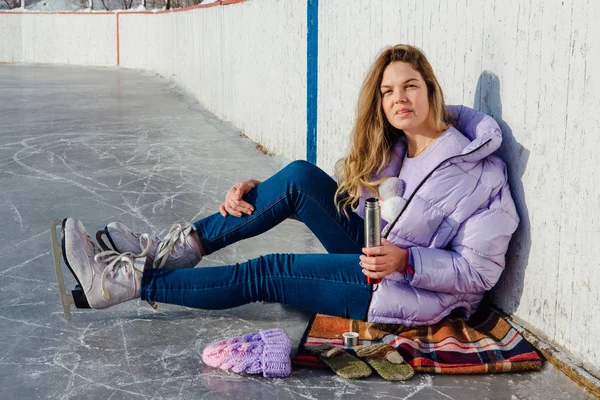 Adorável jovem relaxante depois de andar de patins no gelo e beber bebida quente de termo pot na pista de gelo . — Fotografia de Stock