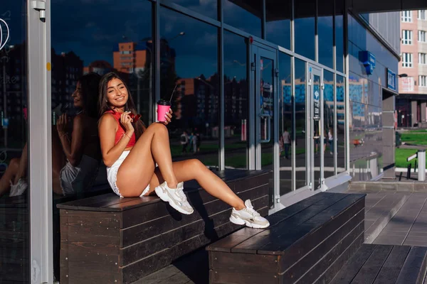 时尚快乐的年轻黑发女人穿着白色短裤和运动鞋拿着粉红色的咖啡杯去坐在咖啡店旁边. — 图库照片