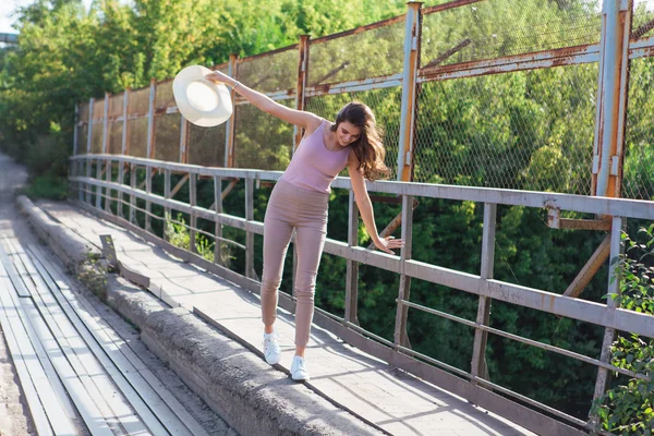 Pretty young woman wearing cowboy hat posing on the old rusty bridge during sunset. — Stock Photo, Image
