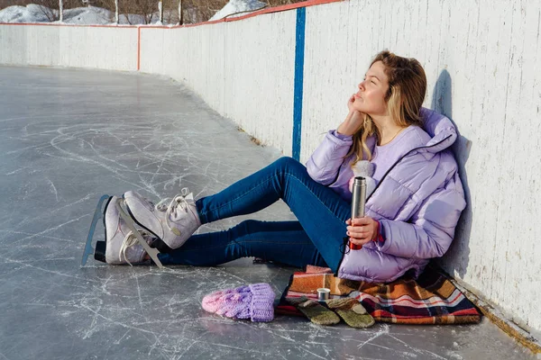 Krásná mladá žena relaxační po jízdě na bruslích a pití teplého nápoje z termo hrnce na kluzišti. — Stock fotografie