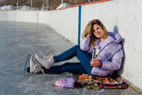 Lovely young woman relaxing after riding ice skates and drinking hot drink from termo pot on the ice rink. — ストック写真