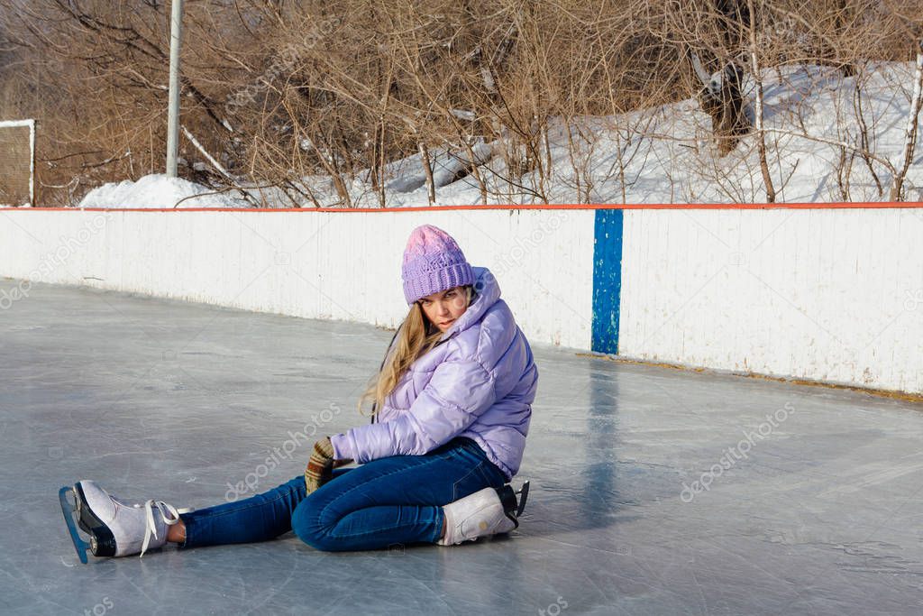 Lovely young woman sitting on the ice rink with ice skates on feet.