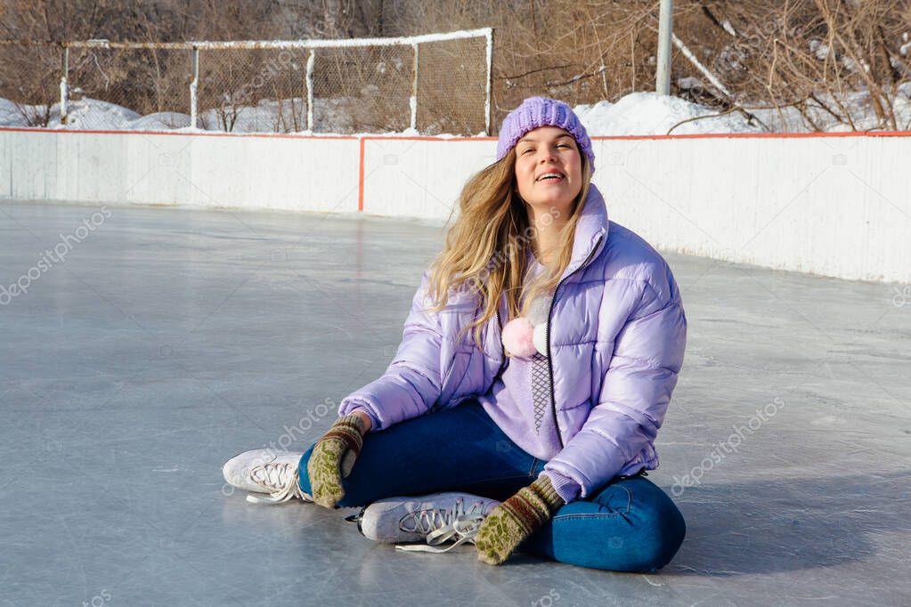 Lovely young woman sitting on the ice rink with ice skates on feet.