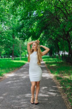 Ağaçların altında duran, güzel beyaz elbiseler giyen çekici sarışın bir kadının portresi..