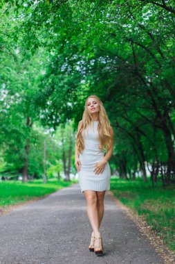 Ağaçların altında duran, güzel beyaz elbiseler giyen çekici sarışın bir kadının portresi..