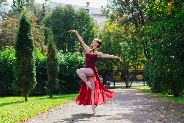 Woman ballerina in red ballet dress dancing in pointe shoes in autumn park.