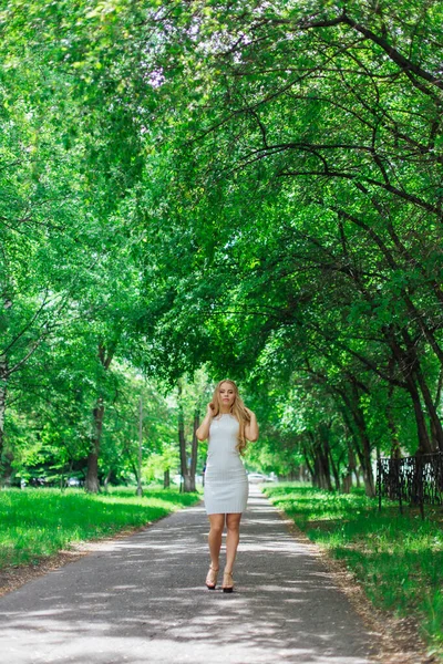 Retrato de uma mulher loira encantadora vestindo belo vestido branco em pé na estrada sob árvores . — Fotografia de Stock