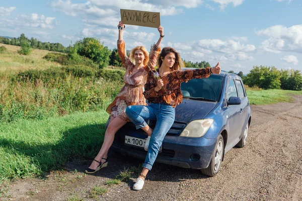 Two beautiful girls hitchhiking and vote with a sign ANYWHERE sitting on the hood of a car on roadside. — Stock Photo, Image